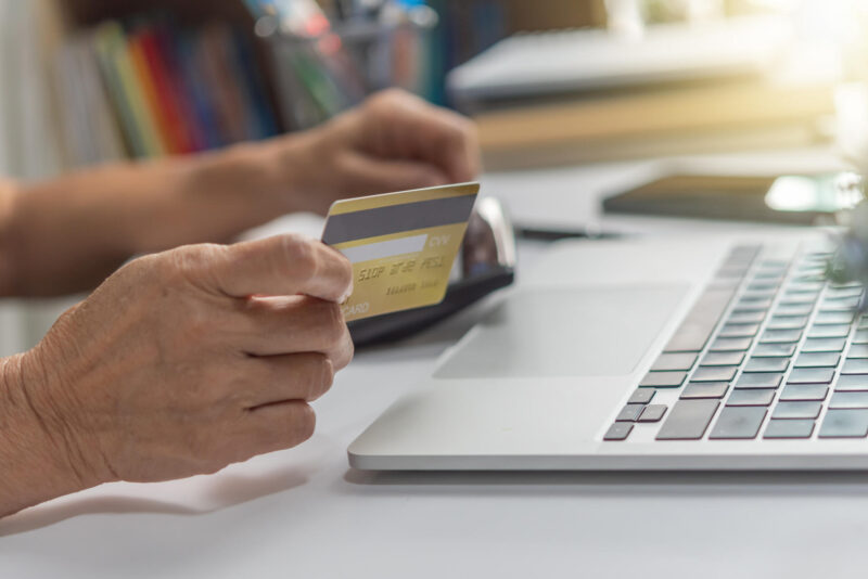 Lähikuvan vanhan ihmisen käsistä ja kannettavasta tietokoneesta. Ihmisellä kädessä pankkikortti.