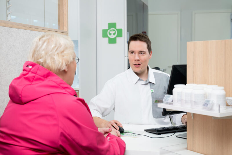 Iäkäs naisasiakas ja apteekin farmaseutti keskustelevat apteekin reseptitiskin ääressä.