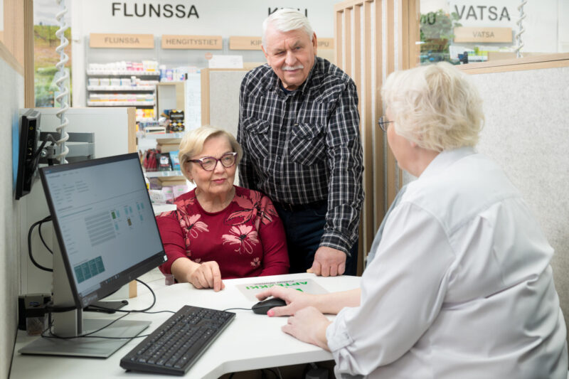 Iäkäs pariskunta apteekin  reseptitiskissä juttelemassa farmaseutin kanssa.